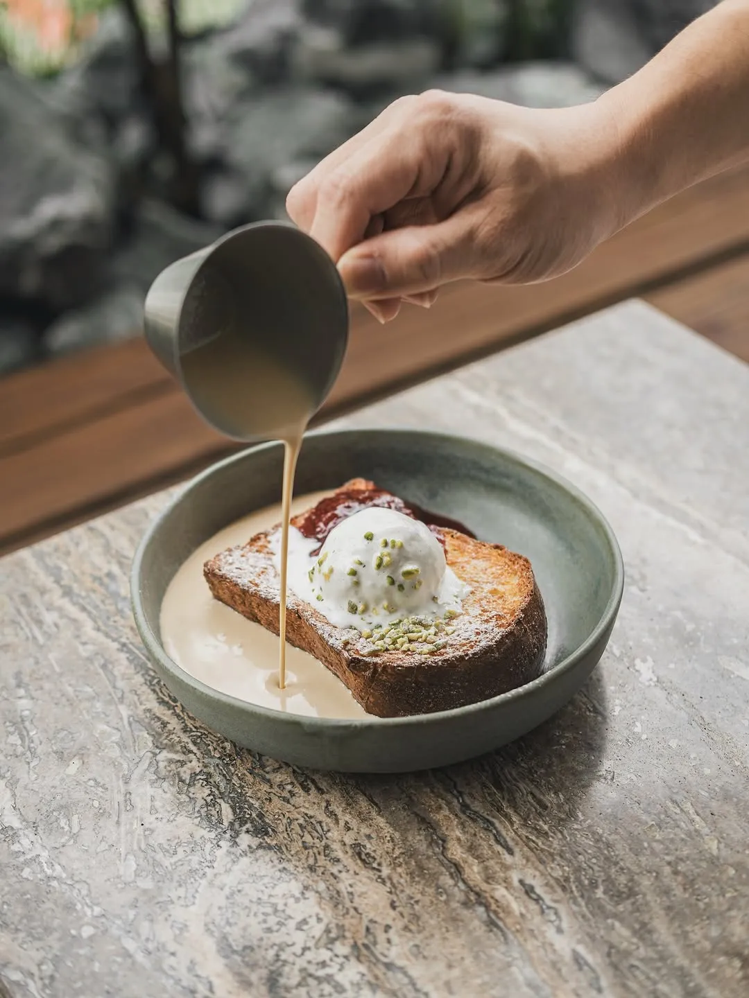 Hand pouring sauce over dessert on a stone surface.