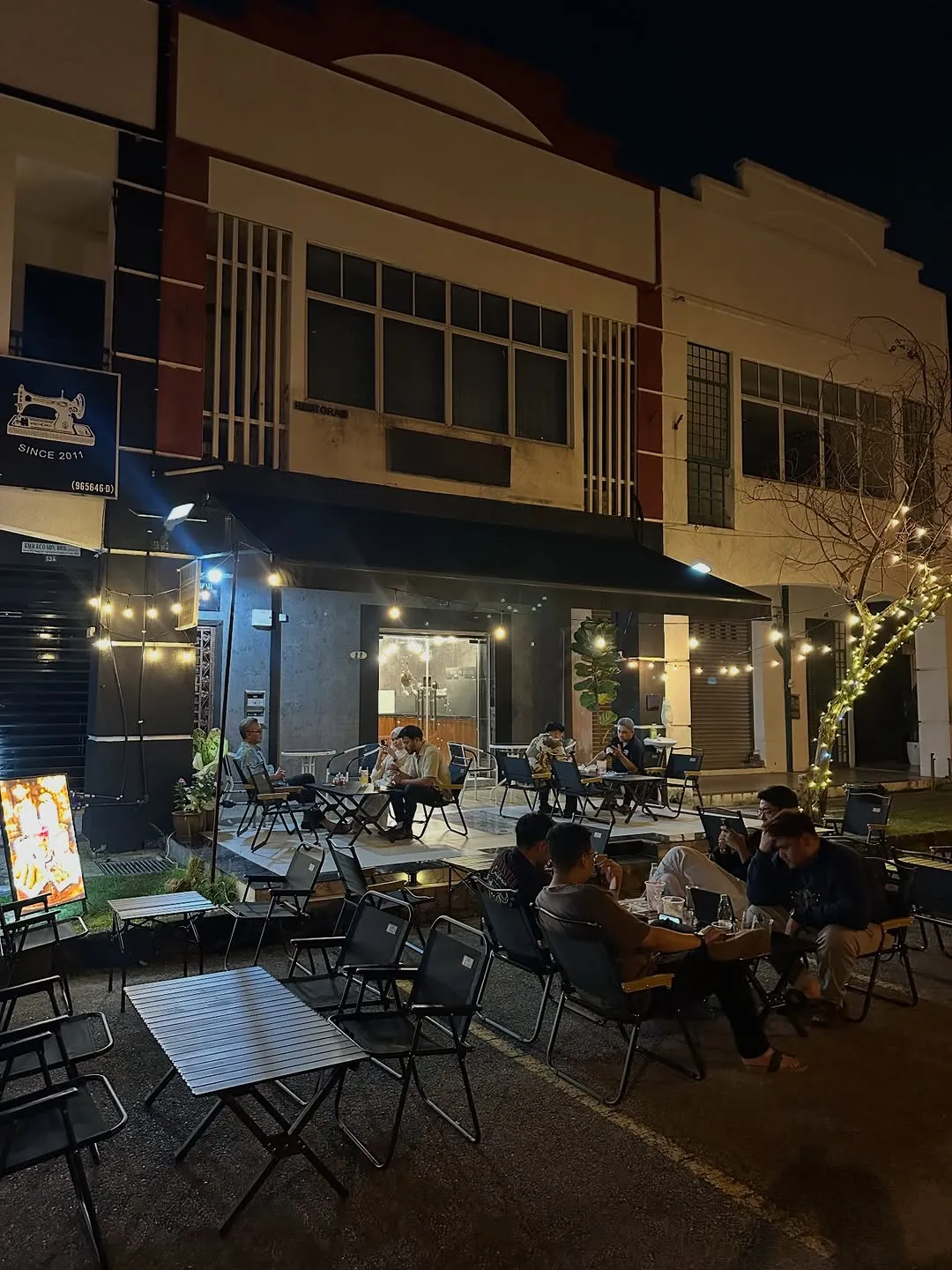 Outdoor seating area with people enjoying drinks under warm lights at night.
