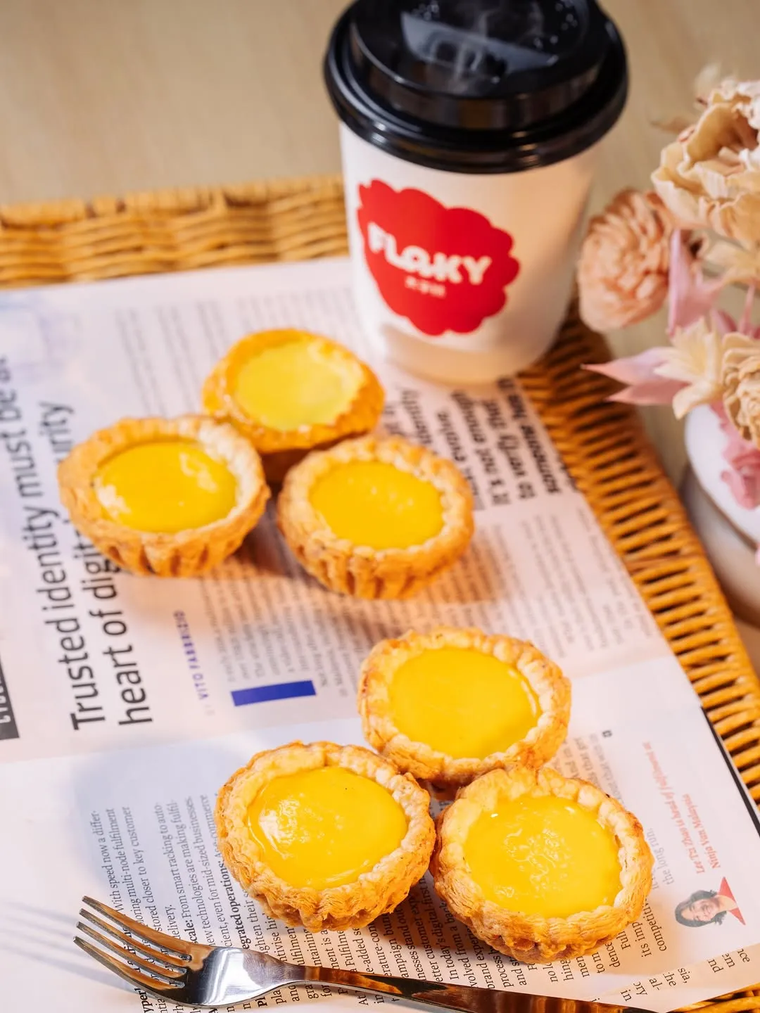Six golden egg tarts on a newspaper with a coffee cup.