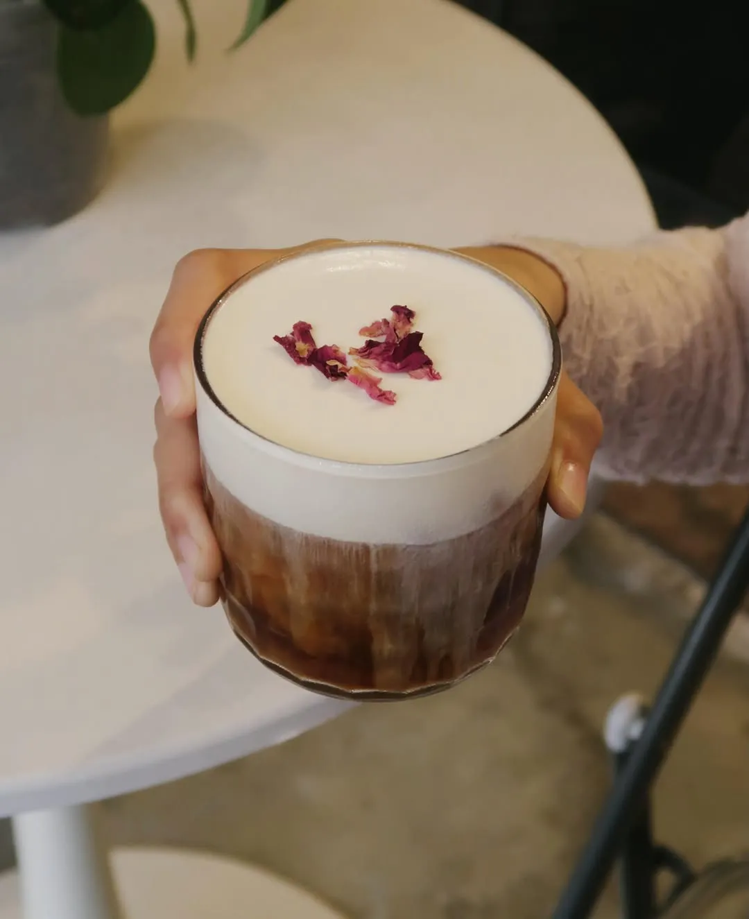 A hand presenting a glass of vanilla cloud cold brew garnished with dried rose petals.