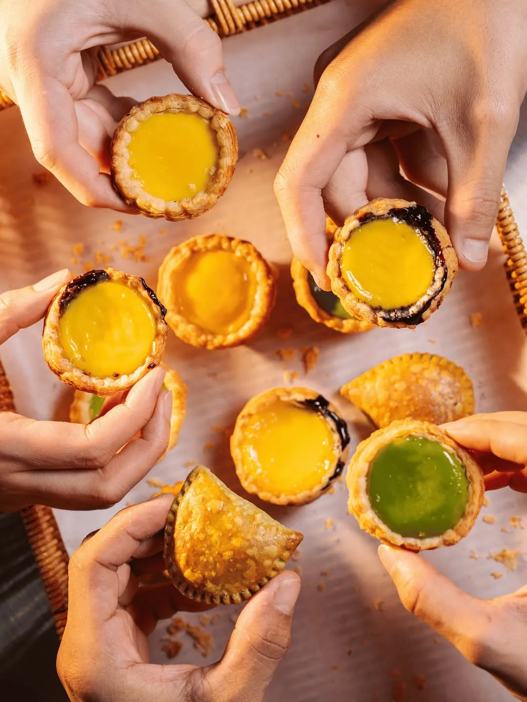 Hands holding colorful egg tarts in a wooden basket.