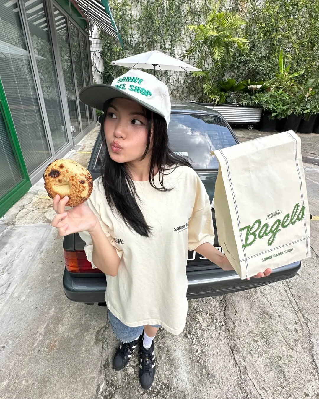 Person holding a bagel and takeout bag outside a cafe with greenery.